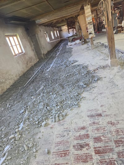 The image shows the interior of a building in Kaleytsa, Bulgaria. The foreground is dominated by a concrete floor partially covered with rubble and a metal grid. The wall to the left has a window with a grid-like design and several smaller windows higher up. The right side of the image reveals a brick pattern in the floor. The background contains wooden beams and various objects, suggesting a workshop or storage area.