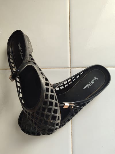 Two black jelly flats with a caged design are placed on a white tiled surface. The shoes are positioned at an angle, with the left shoe overlapping the right. Both shoes have a strap across the instep with a small gold buckle. The brand name "Youth Balance" is visible in white lettering on the insole of each shoe. The tiles are arranged in a grid pattern, with visible grout lines. The lighting suggests an indoor environment, possibly a home.