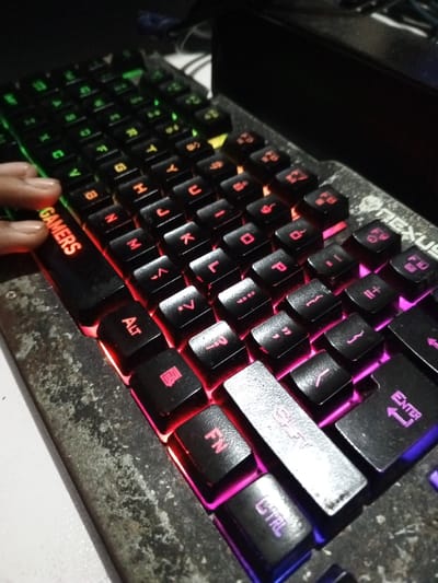 The image displays a close-up, angled perspective of a black mechanical gaming keyboard with multi-zone RGB backlighting, located in an indoor setting within Al Haouafate, Morocco. The keyboard's base exhibits a distinct, heavily textured, mottled grey surface, possibly indicating wear. The brand "NEXUS" is visible on the upper right side of the keyboard chassis. Keycaps are black, with alphanumeric and function legends illuminated. Backlighting gradients include green on the far left keys, transitioning to red/orange for the central alphanumeric keys, and further to pink/purple/blue for the right-side control keys such as 'Shift', 'Enter', 'Fn', and 'Ctrl'. A human hand, specifically the tips of a thumb, index, and middle finger with a light skin tone, is positioned on the left side of the keyboard, resting on or near the 'GMERS' spacebar and adjacent keys. The background is dark and out of focus, revealing a partial view of a black rectangular object and faint cables, consistent with a computer workstation.