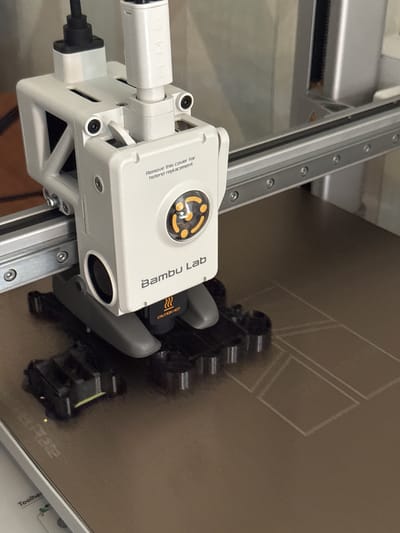 The image shows a close-up of a white 3D printer head, labeled "Bambu Lab," positioned above a textured brown print bed. The print bed has several thin, white lines etched onto it, forming geometric shapes. Attached to the print head is a black component with yellow warning text that reads "CAUTION HOT." The printer is mounted on a silver metal rail system. The background is softly blurred, showing what appears to be a wooden surface and a larger silver structure. There are no people or animals visible in the image. The image does not contain any information that could be used to determine its location in the city of Белоус, Russia.