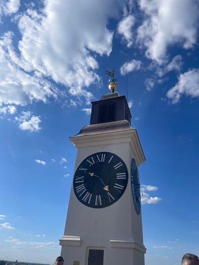 A low-angle shot captures the iconic clock tower of the Petrovaradin Fortress in Serbia, set against a vibrant blue sky dotted with scattered white cumulus clouds. The image is oriented vertically (rotated 90 degrees counter-clockwise), looking upwards at the tower.
The most prominent feature is the large, dark-faced clock with contrasting white Roman numerals (I through XII) and gold-colored hands. Notably, the Petrovaradin clock is famous for its "drunken" feature, where the longer hand indicates the hour and the shorter hand indicates the minutes. In this image, the long hour hand points towards VI (6), and the short minute hand points towards XII (12), indicating the time is approximately 6:00. Above this main clock face, a smaller, partially visible clock face with Roman numerals is also present on the tower.
The tower itself is constructed from light-colored stone or plaster, featuring decorative molding and a darker, louvered roof structure at its apex. Crowning the tower is a golden spherical ornament from which a decorative weather vane, resembling a small flag or banner and a figure, extends upwards. A thin, antenna-like rod also protrudes from the very top.
The weather is clear and sunny, indicative of daytime, likely late afternoon given the clock's reading of 6:00. Partial views of two individuals are visible in the original image's corners; a glimpse of a person's hair in the top right and a person's head wearing glasses in the bottom right, suggesting human presence at or near the tower. The setting emphasizes the architectural grandeur of the fortress's clock tower against a picturesque sky.