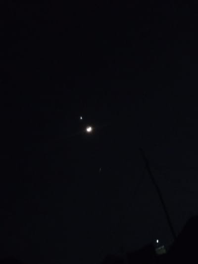 The image is a nighttime photograph of the sky over Ashata, Nigeria. It features a crescent moon and a bright star or planet. There are also faint lights visible in the distance, suggesting an urban environment. A dark, silhouetted object, possibly a tree or building, is present in the lower right corner of the frame. The overall scene is dark, with the celestial bodies and distant lights providing the only illumination.