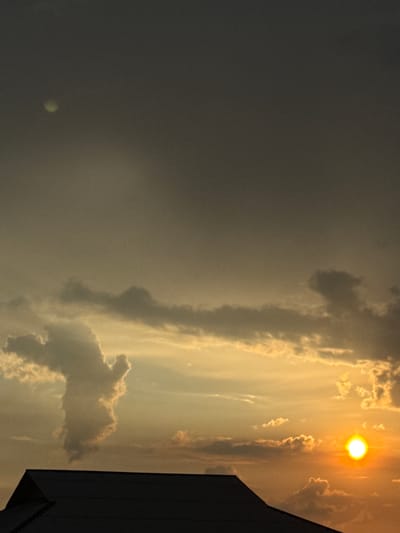 The image depicts a silhouetted rooftop in the foreground against a dramatic sky at sunset. The sky is a gradient of dark grey at the top, transitioning to yellow and orange hues towards the horizon, illuminated by the setting sun. Several clouds are present, some dark and wispy, others bright and illuminated by the sun. A particularly large cloud formation on the left side of the image appears to resemble a human-like figure. The sun is a bright orb located on the right side of the frame, casting a warm glow on the clouds. The scene appears to be an exterior shot, possibly of a rural or suburban area, given the prominent rooftop and the expansive sky. The location in Baş-Bulak, Kyrgyzstan, is a natural landscape setting with a vast sky that is characteristic of the region. There are no people or direct interactions visible in the frame. The overall impression is one of a serene, natural sunset, with the cloud formation adding a unique visual element.