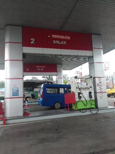 The image depicts a gas station at an unspecified location within Cijedil, Indonesia, under an overcast sky. The foreground shows an asphalt pavement leading to a multi-pump fuel island covered by a large canopy. The canopy's support pillars are white with red accents, and overhead signs display "PERTALITE" and "SOLAR" under numbered bays "2" and "3." A blue minivan is positioned at a fuel pump on the left side of the frame, facing right, suggesting it is either refueling or waiting. The fuel pump itself is primarily white with green and black accents on its lower section, featuring multiple hoses. To the left of the minivan, another pump stand is visible, and a small red shelving unit holds several white bottles. In the background, behind the blue minivan, a white and green passenger bus is partially visible. Further in the distant right background, a yellow truck is present. No individuals are clearly identifiable within the frame.