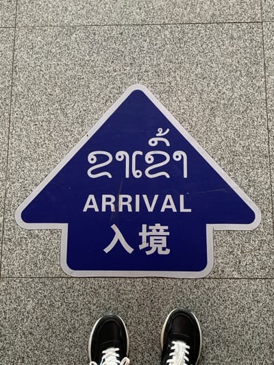 An arrow-shaped blue sign is affixed to a speckled gray tiled floor. The sign contains white text in three languages. The top line features text in what appears to be Lao script. The middle line reads "ARRIVAL" in English. The bottom line displays Chinese characters. Two black and white sneakers are visible at the bottom of the frame, positioned on the tiled floor in front of the sign. The context suggests this is an arrival marker within a public space in the Golden Triangle Special Economic Zone, Laos, likely an airport or transportation hub.