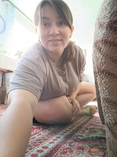 A young woman is captured from a low angle, facing the camera directly and smiling slightly. She has dark hair pulled back into a ponytail and is wearing a plain, light brown t-shirt. Her legs are bent and positioned in front of her, with her left arm resting on her knee. She is seated on a patterned rug with red, blue, and white motifs. To the right of the frame, a large, furry, brown and tan object, possibly a pet or a piece of furniture, occupies a significant portion of the right side. In the background, a wooden table and a window with sheer curtains are visible, with bright light streaming in from the left. A clock can be seen on the wall to the far left. The scene appears to be indoors, likely in a home environment, with no specific indicators pointing to the city of Каменное, Russia.