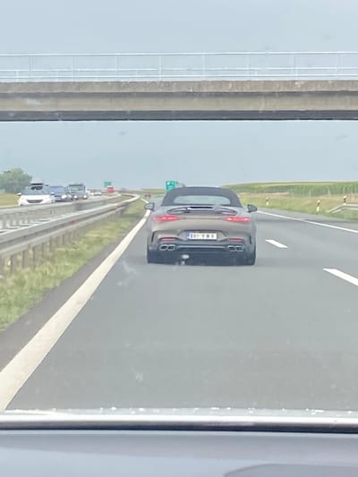 The image shows a grey convertible car driving on a highway in Kovilj, Serbia. The car has a license plate that reads "BGV VMY". In the background, there is a highway overpass and agricultural fields. Other vehicles are visible on the highway, including cars and trucks. The sky is overcast.
