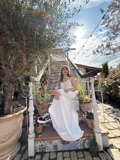 A woman is seated on a tiled outdoor staircase, wearing a long white dress and black sneakers. She has tattoos on her arms and is wearing rings. The staircase is adorned with potted plants, including an olive tree on the left. In the background, there is a building with a tiled roof and blooming pink roses. The sky is partly cloudy, and sunlight is visible. The scene appears to be in a garden or courtyard setting. There is no indication that this location is in Semogorskiy, Russia.