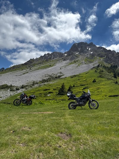 The image depicts a scenic mountain landscape in Jošanica, Montenegro, with two adventure motorcycles parked on a grassy slope. The foreground is dominated by lush green grass dotted with small yellow wildflowers. In the mid-ground, two motorcycles are positioned. The motorcycle on the left is orange and white, equipped with luggage racks. The motorcycle on the right is black and also features luggage. Behind the motorcycles, the terrain ascends into a steep, rocky mountainside covered in patches of green vegetation and scree. The upper portion of the image features a dramatic mountain peak with rugged rock formations and remnants of snow. The sky is a vibrant blue, with scattered white clouds casting shadows on the landscape. The overall scene conveys a sense of exploration and natural beauty.