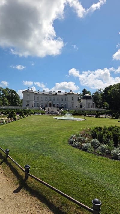 The image shows a stately, light-colored manor house with multiple windows and a mansard roof, set against a bright blue sky with scattered white clouds. The manor is flanked by trees and features a prominent dome-shaped structure on its right side. In front of the manor, a wide, manicured green lawn slopes gently downwards. A rectangular fountain with water spraying upwards is located in the middle of the lawn. The foreground is occupied by a gravel path and a dark metal fence with spherical finials. Flowerbeds with various plants and manicured bushes are visible along the edge of the lawn and closer to the manor. A few indistinct figures are seen on the steps leading up to the manor. The scene is characteristic of Palanga's Amber Museum, formerly known as Tiškevičiai Palace, a significant landmark in the city.