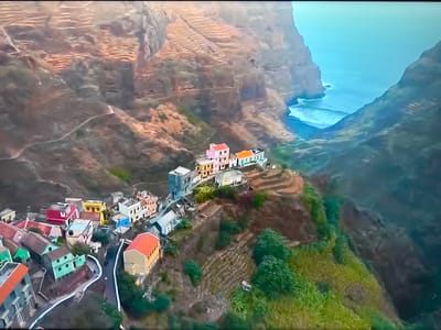 This is an aerial view of a village nestled on steep, terraced hillsides overlooking the ocean. The village consists of numerous brightly colored houses with red, yellow, pink, green, and blue facades, clustered together along winding paths. The surrounding landscape is characterized by rugged, rocky cliffs and lush green vegetation, with cultivated terraces visible on the mountainside. The ocean appears a vibrant blue, with waves crashing against the base of the cliffs. The image depicts a remote, scenic settlement in a dramatic natural environment. There is no indication of the city of Ləcət, Azerbaijan, in this image.