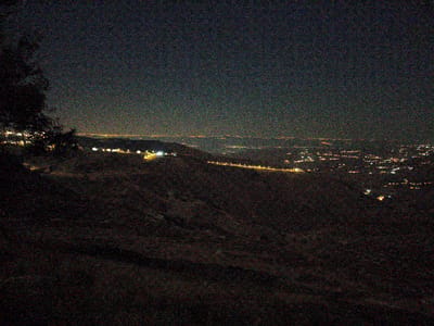 The image depicts a nighttime landscape overlooking a sprawling, illuminated city in the distance, likely in the Umm Al-Qatin region of Jordan. The foreground is dominated by a dark, textured hillside, with a shadowy tree branch visible on the left. Below the tree, a cluster of bright lights suggests a settlement or a section of the city closer to the vantage point.

The main subject of the image is the vast expanse of city lights stretching across the horizon. These lights form a dense tapestry of yellows and whites, outlining roads and buildings, suggesting a significant urban area. A faint, continuous line of lights in the far distance could indicate a highway or a further settlement. The overall impression is one of a bustling urban environment viewed from a high elevation.

The time of day is clearly night, evidenced by the darkness and the prevalence of artificial lights. There are no discernible weather conditions, though the air appears clear enough to allow for a distant view of the city. The location cues are subtle, but the "Umm Al-Qatin, Jordan" context suggests a mountainous or hilly terrain characteristic of this region, which aligns with the elevated perspective in the image. No people or specific objects are distinctly visible, and the scene evokes a sense of quiet observation and perhaps awe at the illuminated urban sprawl. There is no visible text in the image.