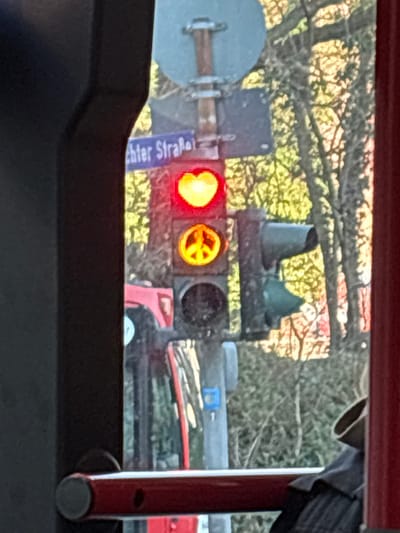 The image presents an outdoor street scene in Aachen, Germany, viewed from an interior perspective, framed by dark and red structural elements. Central to the frame is a vertical traffic light pole. The uppermost signal displays a red heart shape, while the middle signal exhibits a yellow peace symbol. The bottom signal lens is dark and unilluminated. Above this traffic light, a partial blue street sign is visible, with the discernible German text "chter Straße," identifying the location as Richter Straße in Aachen. To the right of the main traffic light, a secondary traffic signal displays a green light. The background consists of blurred, bare tree branches with subtle hints of yellow-green foliage. In the immediate foreground, a red horizontal safety bar with a metallic end cap is visible at the bottom, positioned in front of dark fabric. A portion of a red vehicle is faintly discernible to the lower left, reflecting elements or seen through a window.
