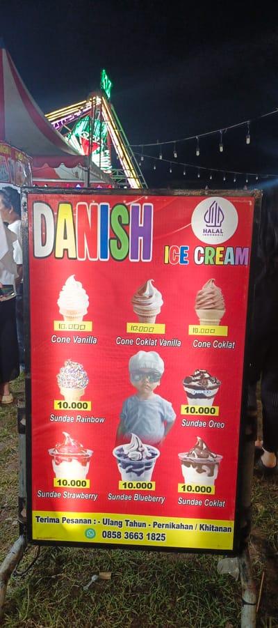 This is a photograph of an outdoor ice cream stand at night, likely in Banding Agung, Indonesia, given the Indonesian text on the sign. The focal point is a large, red banner advertising "DANISH ICE CREAM". The banner displays various ice cream flavors and sundaes with prices listed in Indonesian Rupiah. Prominently featured are images of cones with vanilla and chocolate ice cream, and sundaes with rainbow sprinkles, strawberry, blueberry, and Oreo toppings. The price for most items appears to be 10,000 Rupiah. In the center of the banner, a young child wearing a cap and sunglasses is depicted. At the bottom of the banner, contact information for placing orders is provided, including a phone number and a WhatsApp icon. In the background, elements of a fairground or festival are visible, including colorful lights and tent structures. The ground is covered with grass. The overall scene suggests a lively, festive atmosphere associated with a local event or market.