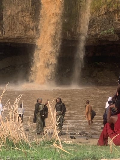 The image shows Gurara Waterfalls in Kukum, Nigeria. The waterfall has muddy water flowing from a rocky cliff into a shallow pool. Several people are visible: one standing alone in the pool, a group standing in the water at the edge of the pool, and two individuals standing on the grass at the edge of the pool, near dried reeds.