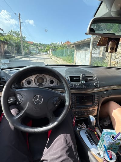 The image shows the interior of a Mercedes-Benz car, likely a W211 model, with the driver's perspective of the road ahead. The car is stationary in what appears to be a residential area. The steering wheel is visible with the Mercedes-Benz logo prominently displayed. The dashboard is also visible, with various controls, vents, and the car's infotainment system. The radio is tuned to 87.5 MHz. The instrument cluster shows the car's gauges. In the front passenger area, there is a box of "Florina Ice Tea" and a can. The driver's seat is occupied, and a person is wearing black pants with red trim on them. Through the windshield, one can see a road, buildings, trees, utility poles, and a clear, sunny sky. Based on the architecture, the location is likely the city of Rila, Bulgaria. There are cigarette butts in an ashtray. A VIP-branded air freshener is attached to the rearview mirror.