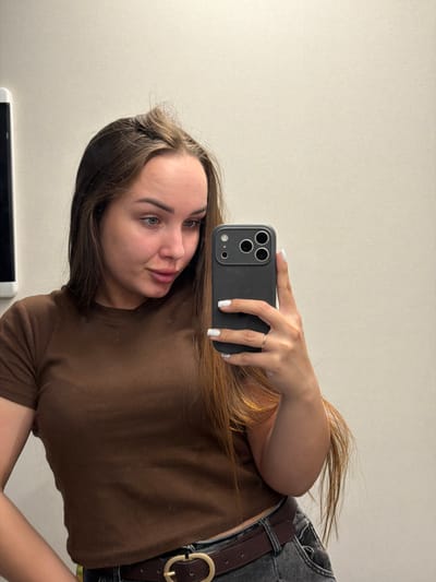 In an indoor setting, a young woman with long, brown hair is taking a selfie in a mirror. She is wearing a brown, ribbed, short-sleeved shirt and dark jeans with a brown belt. Her left hand is resting on her hip, and her right hand is holding a black smartphone up to take the picture. The background is a plain, light-colored wall. There are no discernible landmarks or contextual clues to suggest the location is in Kamyennoye, Russia.