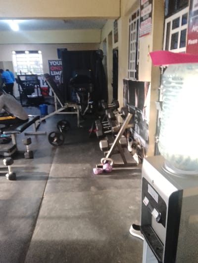 The image depicts the interior of a gym located in Njoro ward, Kenya. The environment features a dark rubber mat floor with visible seams, light-colored walls, and multiple windows with white frames along the far wall. Overhead fluorescent lights illuminate the space.

In the foreground, a silver and black water cooler with a large clear plastic water bottle sits on the right. A pink object rests on the water bottle. A black and white athletic shoe is positioned on the floor next to the cooler's dispenser tray.

Towards the mid-ground, a person wearing a grey shirt is seated on a black-padded weight bench. Two black dumbbells are on the floor to the left of this bench. Directly in front of the water cooler, a silver weight rack holds multiple dumbbells. Two purple kettlebells are on the floor near the base of this rack. A single barbell plate lies on the floor further back. An incline weight bench with black padding is visible behind the seated individual. A poster on the back wall displays partially visible text: "YOU[R] ONLY LIMI[T IS]".

In the background, another individual wearing a blue shirt is partially visible near the far window, standing amongst additional gym equipment, including what appears to be a cardio machine and other weight stations. A blue bucket is on the floor to the left of the cardio machine. The right wall features white-framed elements and partially visible signs, one of which appears to indicate a price such as "Ksh. 2,500." No active interactions are observed between individuals.
