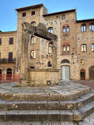 The image captures the Pozzo della Cisterna (Well of the Cistern) located in Piazza della Cisterna, San Gimignano, Italy. The central subject is an ancient stone well, composed of a hexagonal wellhead resting on a multi-tiered, octagonal stone platform. Two robust vertical stone pillars flank the wellhead, supporting a massive horizontal stone beam overhead, which is heavily encrusted with moss and lichen. The immediate ground around the well is paved with large, irregular stone slabs.

Surrounding the well are several multi-story medieval stone buildings, constructed primarily from light brown and grey stone blocks with brick accents, particularly around arched windows and doorways. These buildings feature numerous windows, many with arched tops; some have closed brown wooden shutters, while others contain potted plants or show reflections of the sky and adjacent structures. Tiled roofs are visible at the top of the structures. On the far left, a building displays a wooden balcony. An area to the left of the well is demarcated by orange construction barriers, partially obscuring an arched entrance displaying "LE TERRAZZE". Overhead, multiple strings of decorative lights crisscross the clear blue sky between the buildings. The scene is illuminated by natural light, creating distinct shadows on the stone surfaces. No individuals are present in the image.