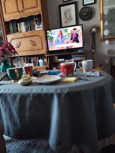 This image captures a cozy living room scene, likely in Poland, with a round table covered in a grey tablecloth as the central focus. On the table, various objects suggest a relaxed morning. Two mugs, one red with a white winter tree pattern and another plain white, sit next to a pack of cigarettes. A small, decorative ceramic pot with a lid is also present, alongside a green mug with a gold handle, and a small white plate with a floral design. A yellow sticky note and a black pen are laid out, perhaps for jotting down notes.

Behind the table, a television displays a talk show with two female hosts. One host, in a blue suit, is being interviewed by another in a pink blazer. The time "09:07" is visible on the screen, indicating it's a morning broadcast. The text on the screen translates to "Can fear of flying be overcome?" and "Smoking kills - quit now!", suggesting the topic of the show and a public health message.

The background reveals a wooden hutch filled with books and decorative items, including a small blue house figurine and a vase of red flowers. To the right, a black circular clock on the wall shows the time is approximately 2:30, contrasting with the time on the TV. A gold-framed mirror with snowflake decorations hangs on the wall, adding a touch of festive charm. The overall atmosphere is one of domestic comfort, with the gentle morning light suggesting a quiet start to the day.