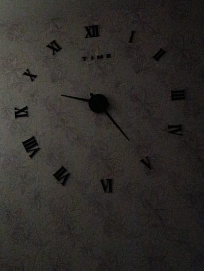 The image shows a wall clock with Roman numerals on a patterned wallpaper. The clock hands are visible, indicating a time that is difficult to determine precisely due to the low lighting and shadows. The Roman numerals present are X, XI, XII, I, II, III, IV, V, VI, VII, and VIII. The word "TIME" is also visible on the clock face. The wallpaper has a subtle floral or abstract pattern. The lighting is dim, with shadows obscuring parts of the clock and the wallpaper. There is no information to suggest this image is located in Verkh-Chumanka, Russia; it is a general depiction of a wall clock.