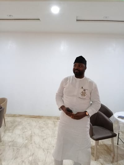 The image shows a man in a white long-sleeved shirt and black cap standing in a room. He is holding a black cell phone in his right hand, and his left hand is resting on his right wrist. There are two chairs and a table with an object on it to his right. The room has white walls, tiled floors, and a white ceiling with recessed lighting. The man is looking towards the left of the frame. The image is taken from a perspective that appears to be slightly below the man's eye level. The man is the only person in the image. The location is an interior space in Yola, Nigeria (coordinates: 9.198, 12.492). It is most likely an office or waiting area.  The setting is neutral, and there are no other objects that indicate the time of day or the purpose of the room.