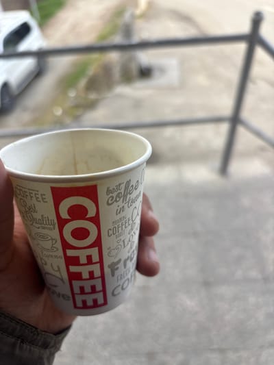 The scene captures a close-up, downward-angled perspective of a human hand holding a white disposable paper coffee cup. The cup features a prominent vertical red bar with "COFFEE" in white capital letters on one side, and various coffee-related words and icons in grey text on the remainder of its surface, including phrases like "best coffee in town" and "morning coffee." The cup's interior shows residue consistent with a consumed beverage, indicating it is partially empty. The hand holding the cup displays visible skin texture and creases. The background is significantly blurred, depicting an outdoor environment. Immediately behind the cup, a dark, thin metal railing runs horizontally across the frame. Below and beyond the railing, a light-colored concrete or paved surface is visible, alongside which a portion of a white passenger vehicle is discernible on the left. Faint green foliage or grass appears next to the paved area in the distance. The overall impression is an individual holding a coffee cup outdoors from an elevated position. This scene is located in the city of Lehvaz, Armenia.