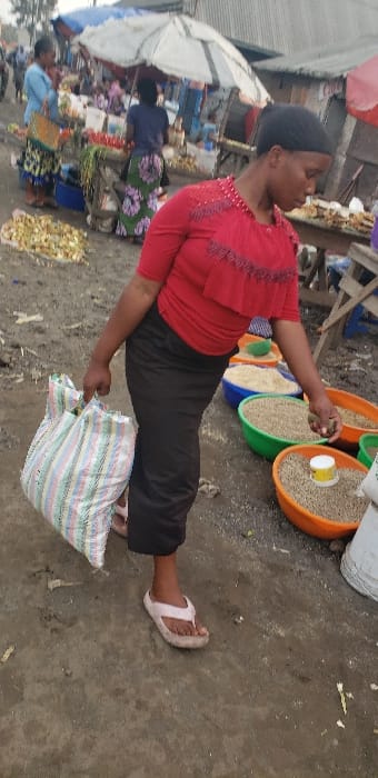 The image captures an outdoor market scene in Buzove, Democratic Republic of the Congo. The central figure is a dark-skinned female, positioned facing slightly right, wearing a red short-sleeved top with ruffled and beaded embellishments, dark long trousers or a skirt, and light-colored flip-flops. Her left hand grasps a large, multi-colored striped woven bag (white, green, pink, blue, beige). Her right hand is lowered towards a series of large, open, colorful plastic basins (orange, green, blue) arranged on the unpaved ground to her right, each filled with dry granular goods resembling grains or seeds. A small white and yellow container rests within one orange basin.

In the midground, other market activities are visible. To the left of the main subject, another female vendor, seen from behind, wears a green patterned skirt and stands behind a stall displaying produce, including red items resembling tomatoes. Further left, a third female in a light blue top and patterned skirt is partially visible. The background consists of rudimentary market stalls with wooden frames and corrugated metal roofs or walls, and large white and red umbrellas providing shade over various merchandise. The ground is unpaved, appearing as compacted earth with scattered debris and areas of dampness. Numerous other individuals are present in the blurred background, indicating a busy public space.