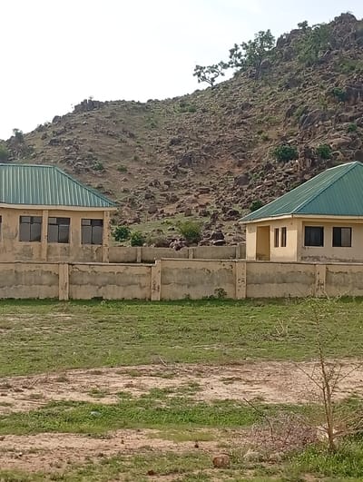 The image depicts a residential or institutional area with two light-colored buildings featuring green, corrugated metal roofs. A low, cream-colored concrete wall runs horizontally across the mid-ground, separating the buildings from a grassy foreground. The buildings appear to be single-story structures with dark-framed windows. The left building has a series of windows, while the right building has fewer windows and a visible entrance. Behind the buildings, a rocky, arid hillside dominates the background, dotted with sparse vegetation and a few trees. The sky is overcast, suggesting a bright but diffused light. The foreground consists of a patch of dry, uneven ground with some patches of green grass. The overall impression is of a sparsely populated, possibly rural or semi-rural environment, consistent with the town of Shuwa, Nigeria, known for its rocky terrain.