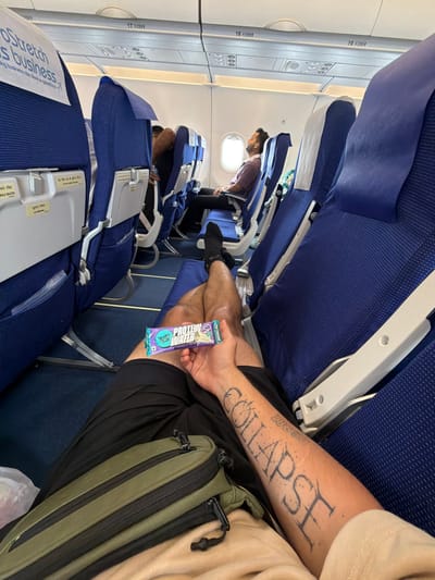An overhead perspective shot captures the interior of an airplane cabin, focusing on a passenger's legs and arm. The passenger, wearing black shorts and a light-colored t-shirt, is holding a "Yoga Bar Protein Wafer" in their right hand. The left arm, resting on a tan blanket, displays a large tattoo that reads "COLLAPSE" and another word that is partially obscured. A green and black fanny pack is positioned across the passenger's lap. The airplane seats are upholstered in blue fabric, with white headrests. Another passenger is visible in the background, sleeping in a reclined seat. The overall environment is indoors, on a commercial aircraft. The location within the city of Mohan, India, cannot be determined from the image.