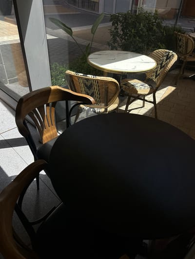 The image depicts an outdoor seating area, likely a cafe or restaurant patio, in Rehovot, Israel. The scene is bathed in sunlight, casting strong shadows. In the foreground, a large, dark, round table dominates the frame. To the left and partially visible are two wooden chairs with curved backs and legs. Behind these chairs and the dark table, a smaller round table with a white marble top and gold trim is visible, accompanied by two chairs. These chairs have a woven, rattan-like texture with black and white patterns on the seats and backs, and appear to be of a French bistro style. A leafy green plant grows behind the smaller table. The flooring is a mix of light-colored tiled pavement and darker, rougher paved bricks. In the background, through glass or a clear partition, a sidewalk and possibly more seating areas of the establishment can be glimpsed, along with vegetation and structures that suggest an urban environment within Rehovot. No people are present in the immediate scene. The overall impression is one of an unoccupied, sunlit outdoor dining space.