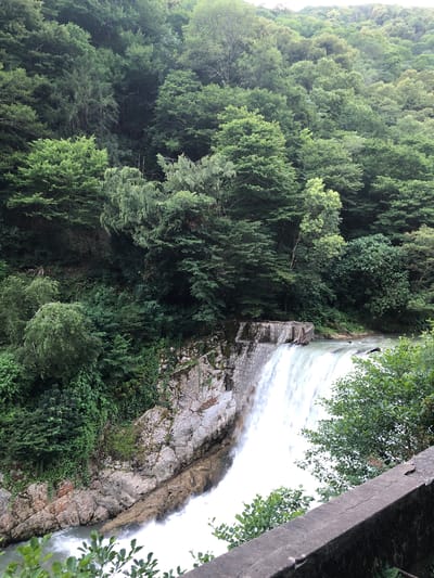 This media file captures a scenic view of a waterfall cascading over a rocky embankment, surrounded by lush green trees. The perspective is from above, looking down at the falls. The water appears white and frothy as it tumbles down, suggesting a strong current. The rocks are a mix of grey and brown, with patches of moss and vegetation growing on them. The trees are dense and form a thick canopy, creating a natural, verdant setting.
To the right, a portion of a concrete structure is visible, possibly part of a dam or bridge. The sky is overcast, contributing to a soft, diffused light that illuminates the scene. The overall atmosphere is serene and natural, highlighting the power and beauty of the waterfall within its forested environment. The image appears to be taken in a natural, possibly mountainous region given the presence of a waterfall and dense forestation. No people or text are visible in the image.