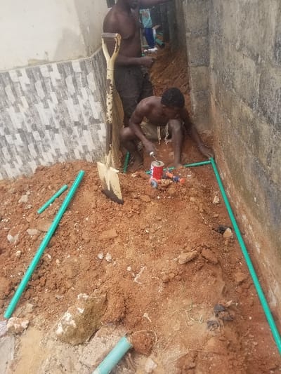The image depicts an outdoor plumbing installation in Edu Town, Nigeria. Two shirtless adult males are present in a narrow, excavated trench with reddish-brown soil.
In the foreground, a dark-skinned male crouches, focused on connecting or treating bright green plastic pipes laid in the dirt. He is positioned near a red and white can (possibly adhesive or primer) and several small blue and orange tools or fittings.
Behind him and slightly to the left, a second dark-skinned, shirtless male stands, observing or engaged in work further down the trench. A large, yellow-handled spade is stuck upright in the soil between the two men.
The environment includes a wall with grey and white patterned tiles to the left and a rough, grey concrete block wall to the right. Additional sections of green plastic piping and a piece of light blue pipe are scattered in the excavated earth. A third individual is partially visible in the distant background.