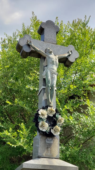 A close-up, low-angle shot captures a weathered statue of Jesus Christ on a granite cross, located in Vyšné Nemecké, Slovakia. The statue, appearing to be made of bronze, depicts Jesus with arms outstretched, wearing a loincloth. The cross, made of speckled grey granite, has ornate clover-shaped ends and bears the inscription "INRI" at the top. A wreath of artificial light-colored flowers and dark greenery adorns the base of the statue, just above a carved chalice symbol. The background is dominated by lush green foliage from trees and a hint of a building on the right. The sky above is partly cloudy. The statue shows signs of age, with peeling paint or patina on the figure of Jesus, and the granite appears slightly weathered.