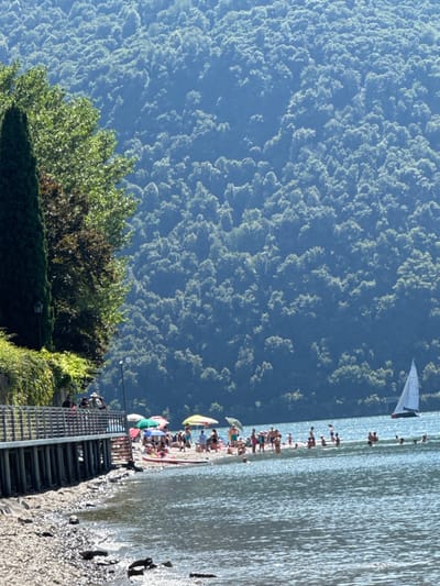 The image presents a sunlit, crowded pebble beach scene on Lake Como, specifically in Abbadia Lariana, Italy. In the foreground, calm, reflective lake water transitions to a narrow, rocky shoreline. Extending from the left is a raised wooden pier or walkway with metal railings, supported by vertical posts, and bordered by dense green foliage, including a tall, dark conical tree. A streetlamp is visible next to the walkway.
The midground features a busy beach area densely populated with individuals of varying ages, including adults and children. Numerous people are standing, sitting, or lying on the sand, with several colorful beach umbrellas providing shade (including yellow, blue, and red umbrellas). A red and white paddleboard or small boat rests on the beach near the water's edge. Many individuals are observed wading or swimming in the shallow, clear lake water adjacent to the beach. Further out on the lake, to the right of the beach, a white-sailed sailboat moves across the water.
The background is dominated by a large, steep mountain face densely covered in green trees and foliage, appearing misty in the distance under a clear, bright sky.