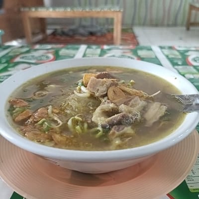 A close-up shot presents a bowl of Indonesian beef soup, "Soto," sitting on a table in Cangkring, Indonesia. The bowl is white, cradled on a peach-colored saucer. The soup is filled with visible pieces of beef, bean sprouts, and fried onions. The table is covered with a green and red checkered tablecloth. In the blurred background, a wooden bench is visible. A spoon rests in the soup. Part of a nutrition label is visible next to the bowl.