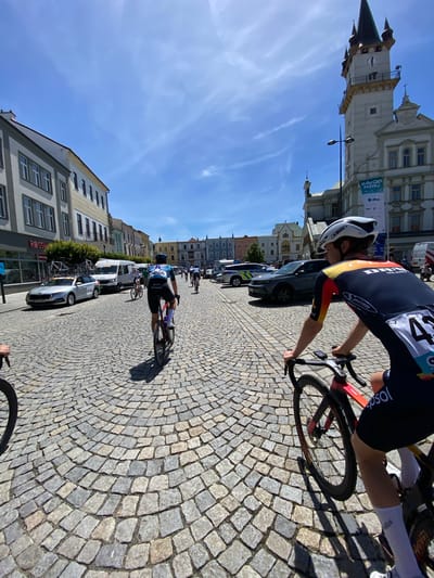 The image captures a cycling race occurring on a cobblestone street in the city of Uničov, Czechia. Cyclists are riding along the street lined with buildings and parked cars. A building with a clock tower is in the background. The sky is blue with some wispy clouds. The foreground shows a cyclist wearing a black, red, and yellow jersey with a white helmet. Further down the street, another cyclist can be seen wearing a blue and white jersey. There are several parked cars and support vehicles lining the street, along with a group of cyclists further down the road.