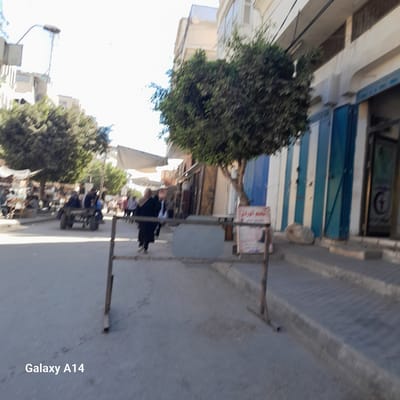 In Deir el-Balah, Palestinian Territories, a street scene unfolds with buildings lining the left side of the frame, featuring a lamppost and balconies. On the right, a building with blue and white striped shutters dominates the view. Several trees are scattered throughout the scene, with one large tree centrally located. People are visible walking and seated in a cart on the street. A metal barrier is present in the foreground, and a sign with Arabic text is affixed to it. The overall environment appears to be a bustling street with commercial activity.