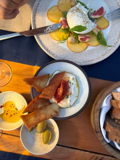 A close-up, overhead shot shows a dining table with various dishes and a person's hand. In the upper portion of the image, a white plate contains burrata cheese, sliced figs, and peaches. A knife with a serrated edge rests on the plate. Below this, a dark-rimmed bowl holds a creamy dip topped with a slice of tomato and what appear to be baked pita chips and olives. To the left of this bowl, a small white dish contains a yellow liquid with red pepper flakes. To the right, a wooden bowl lined with white cloth holds pieces of bread. The table surface is made of wood, and a dark blue tablecloth is visible in the background. The lighting suggests an outdoor setting, possibly a restaurant terrace in Municipal Unit of Sfakiates, Greece, given the rustic wooden elements and the style of the dishes. The overall scene depicts a meal in progress, with focus on the food.
