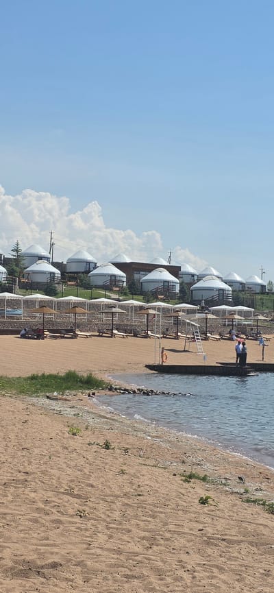 This is a vertical, eye-level, outdoor, full shot of a resort area on the shore of Issyk-Kul lake in Sary-Oy, Kyrgyzstan. In the background, there are numerous white yurt-style accommodations arranged on a grassy slope. In the middle ground, there is a sandy beach with rows of lounge chairs and umbrellas, with a low stone wall separating the beach from the yurts. In the foreground, the sandy beach slopes down to the water's edge, with the light blue water of the lake lapping at the shore. A small wooden dock extends into the water on the right side of the image, with several people visible near it. The sky is a clear blue with some scattered white clouds.