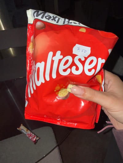 The image captures an indoor scene, reportedly from Tielrode, Belgium, though no specific geographical markers within the city are visually discernible. The primary subject is a large, red "Malteser" brand chocolate candy bag, held by a human hand in the lower right quadrant of the frame. The bag displays "Maxi Pack" at the top and has a white sticker with the handwritten price "7,50" on its upper right side. The hand exhibits manicured nails, with the visible index finger showing a pale, natural tone and a yellow pattern at the tip. Below and to the left of the Malteser bag, a partially unwrapped white chocolate Kit Kat bar rests on a dark, reflective surface. A black cable runs diagonally across this surface, partially obscuring the Kit Kat. The background is uniformly dark and out of focus, indicating low light conditions within an enclosed space.