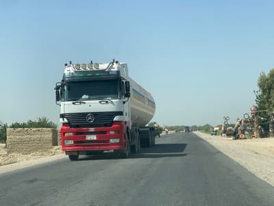 The image shows a white and red tanker truck driving on a paved road in Panjwai, Afghanistan. The truck is traveling in the direction of the viewer and is situated in the left-hand lane of the two-lane road. The road is slightly curved and the tanker truck is the only vehicle visible in the frame. To the right of the truck is a dirt shoulder, where a small group of people is visible. There are several objects on the shoulder, including a blue car and a pile of tires. The road is clear and the sky is bright blue with no clouds. There are small trees and bushes in the background.  A sign on the back of the truck reads "GAS". The road is in good condition with no potholes or cracks.  The truck's license plate number is "1463". The image is taken from a vehicle traveling in the opposite direction to the tanker truck. The photo was taken on a sunny day with clear visibility.  The image was taken on a road leading into the city of Kandahar.