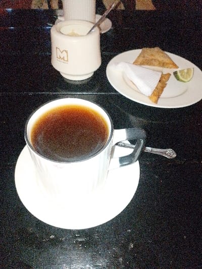 A cup of black coffee sits on a white saucer, accompanied by a teaspoon placed on the table. Behind the coffee cup, there's a sugar bowl with a spoon. A white plate contains a triangular pastry, a piece of food wrapped in a white napkin, and a slice of lime. All of these items are positioned on a black surface that appears to be a table.