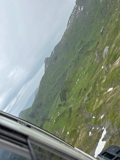 The image shows a panoramic view of a steep, grassy mountainside in Hasliberg Wasserwendi, Switzerland. The mountain is dotted with small white buildings, likely chalets or farmhouses, and a winding path can be seen snaking its way up the slope. Patches of snow are visible on the upper parts of the mountain, contrasting with the vibrant green grass. In the foreground, the edge of a window frame is visible, suggesting the photograph was taken from inside a vehicle, possibly a cable car or train, looking out at the landscape. The sky is overcast with light clouds.
