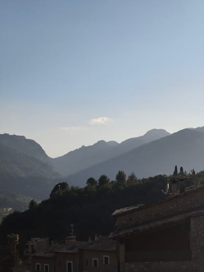 The image depicts a panoramic view of Fornalutx, Spain, characterized by mountainous terrain and traditional architecture, likely during early morning or late afternoon given the lighting.
The upper two-thirds of the frame are dominated by a clear, light blue sky with a subtle atmospheric haze. A single, small, wispy white cloud is present in the upper-mid-left. Below the sky, several layers of mountains extend across the horizon, exhibiting atmospheric perspective with distant peaks appearing as increasingly lighter shades of blue-gray. A distinct diagonal band of sunlight illuminates a section of the mountains in the mid-ground, casting part of the range into shadow.
In the lower portion of the image, a dark green, tree-covered hillside forms a strong horizontal line, separating the distant mountains from the foreground village. The trees appear densely packed, possibly conifers.
The immediate foreground reveals the upper sections and roofs of several stone-built structures typical of the region. The prominent building on the right features a stone facade and a tiled roof, equipped with a chimney and a circular satellite dish. Another chimney is visible further left on the same roofline, accompanied by a small antenna. To the left, additional tiled roofs and partial stone walls of other buildings are visible, including one with a more complex antenna structure. The foreground structures are largely in shadow, contrasting with the brighter sky and sunlit mountain sections. No individuals are visible.