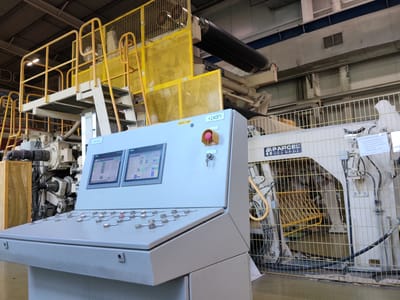 The image shows an industrial setting, likely a factory floor in Shklov, Belarus. The foreground is dominated by a light gray control panel with two screens displaying data and numerous buttons. One screen shows a Siemens logo. A red emergency stop button is prominently visible on the control panel. Behind the control panel, industrial machinery is present, including large rollers and piping, protected by yellow and white safety cages and railings. A large black roller is visible at the top right of the frame. The overall impression is one of a functional manufacturing environment. Some visible text includes "+240P1" and "НАКАТЧИК" on the control panel. On the machinery, "PARCEL CZECH REPUBLIC" is visible. Other text on signs and labels is too small to read clearly. The lighting suggests an indoor setting with artificial illumination. No people are visible in the image.