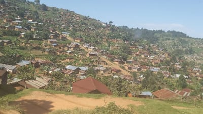 A densely populated hillside settlement comprises small, earthen dwellings with corrugated iron roofs. These structures cascade down the slopes interspersed with lush greenery, likely vegetation and trees. The landscape is uneven, marked by trails and paths. The sky is clear, suggesting a bright, sunny day. Evidence of potential smoke or haze is visible in the distance. The scene captures a rural, possibly developing region.