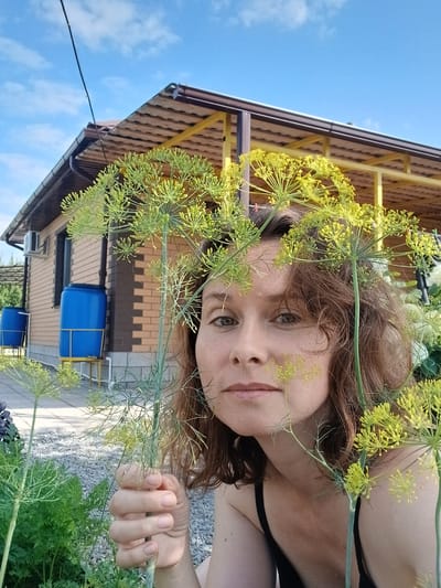 This is a close-up, vertically oriented outdoor photograph of a woman's face partially obscured by dill flowers. The woman has brown hair and is looking towards the camera with a neutral expression. Her face is framed by the feathery green dill blossoms, some of which are yellow. She is wearing a black garment.
In the background, a portion of a building with a brown tiled roof and yellow support beams is visible. Below the roofline, there are blue barrels, possibly water tanks, situated on a paved area. A white air conditioning unit is also mounted on the building. The scene is set against a bright blue sky with scattered white clouds, suggesting a sunny day. The overall impression is one of a casual, perhaps rural, setting. The image appears to have been taken in the daytime. No visible text is present. The photograph was taken in Maslova Pristan', Russia.