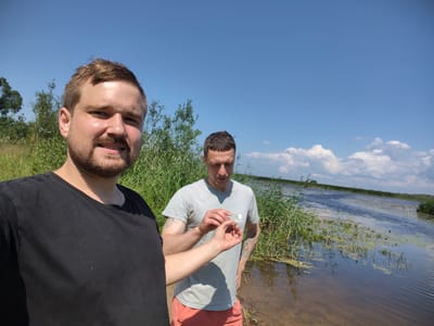 Two men stand outdoors on a sunny day with a body of water and reeds in the background. The man in the foreground, on the left, is wearing a black t-shirt and is looking towards the camera. He has light brown hair and a beard. The man in the background, on the right, is wearing a grey t-shirt and salmon-colored shorts. He is looking down at something he is holding between his fingers, which appears to be a cigarette. He has short brown hair and tattoos on his arms. The environment appears to be a rural area with a river or large pond, surrounded by tall grasses and trees. The sky is blue with scattered white clouds. The location is identified as Malaya Listovka, Russia.