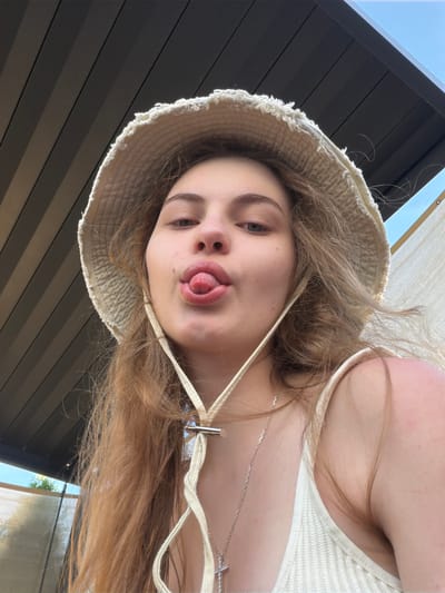 A young woman with long, blonde hair and a bucket hat is pictured from a low angle, looking at the camera with her tongue sticking out. She is wearing a light-colored, ribbed top and a silver necklace with a cross pendant. The background features a dark, corrugated ceiling and a glimpse of a blue sky. The lighting suggests it is daytime. The overall mood appears playful and casual.
